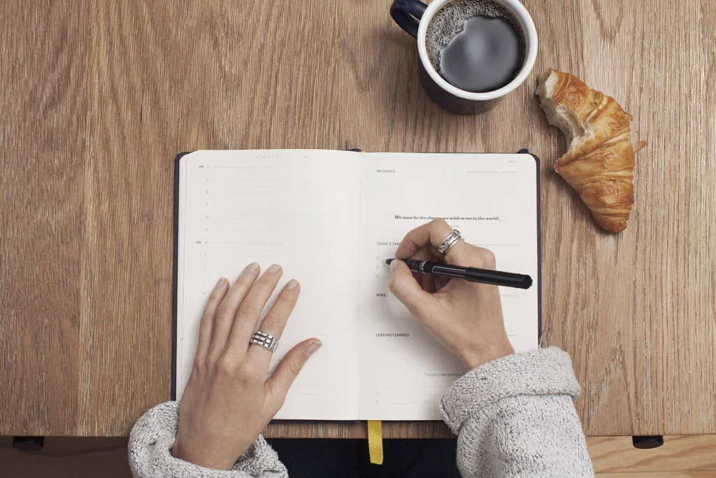 Hands writing in an open journal beside coffee and a croissant on a wooden table