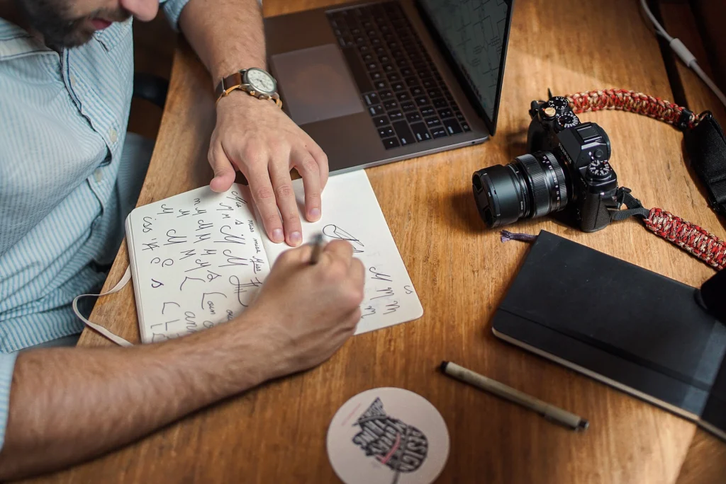 Headless CMS: Person sketching ideas in a notebook next to a laptop and camera