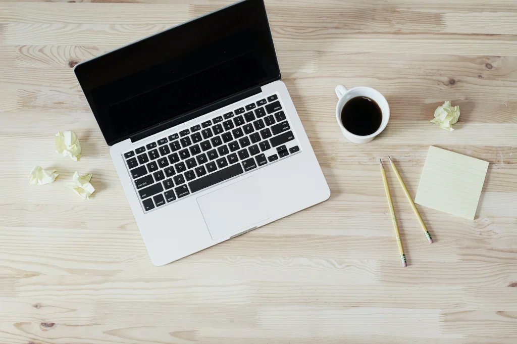 Open laptop, coffee cup and notepad arranged on a pale wooden desk