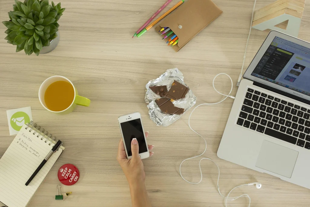 Top view of a desk with a smartphone in hand, a laptop and notebooks