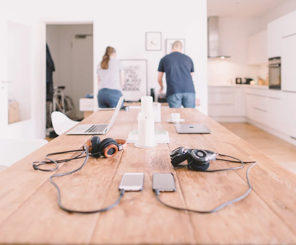 Shared wooden work table with laptops, phones and headphones in a bright office